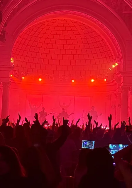 A band on stage under red light in a domed hall in Tucson, the crowd's hands in the air